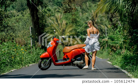 Woman on red scooter in white clothes drive on forest road trail. Dancing trip. One girl caucasian tourist in sunglasses dance, relax near motorbike. Woman on red scooter in white clothes drive on forest road trail. Dancing trip. One girl caucasian tourist in sunglasses dance, relax near motorbike. 105612474