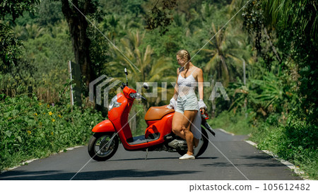 Woman on red scooter in white clothes drive on forest road trail. Dancing trip. One girl caucasian tourist in sunglasses dance, relax near motorbike. 105612482