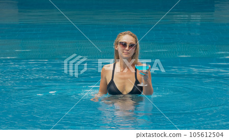 The wide middle shot pretty woman drinking blue cocktail alcohol liquor in swimming pool at hotel. Portrait of sexy girl in pool outdoor. The wide middle shot pretty woman drinking blue cocktail alcohol liquor in swimming pool at hotel. Portrait of sexy girl in pool outdoor. 105612504