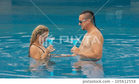 The loving couple hugs and kisses, drinking blue cocktail alcohol liquor in swimming pool at hotel outdoor. Portrait of caucasian man and woman. 105612558