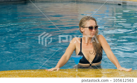 The wide middle shot pretty woman drinking blue cocktail alcohol liquor in swimming pool at hotel. Portrait of sexy girl in pool outdoor. The wide middle shot pretty woman drinking blue cocktail alcohol liquor in swimming pool at hotel. Portrait of sexy girl in pool outdoor. 105612563