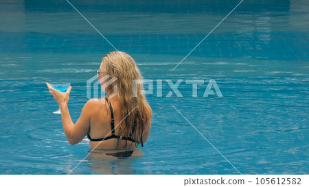 The wide middle shot pretty woman drinking blue cocktail alcohol liquor in swimming pool at hotel. Portrait of sexy girl in pool outdoor. The wide middle shot pretty woman drinking blue cocktail alcohol liquor in swimming pool at hotel. Portrait of sexy girl in pool outdoor. 105612582