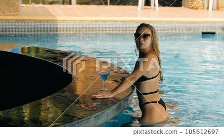 The pretty woman drinking blue cocktail alcohol liquor and sit at the bar. Cocktail bar in the swimming pool. Portrait of sexy girl in pool outdoor. 105612697