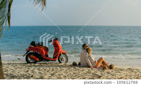 Scooter road trip. Lovely couple on red motorbike in white clothes on sand beach. People walking near the tropical palm trees, sea. Motorcycle rent. Scooter road trip. Lovely couple on red motorbike in white clothes on sand beach. People walking near the tropical palm trees, sea. Motorcycle rent. 105612708