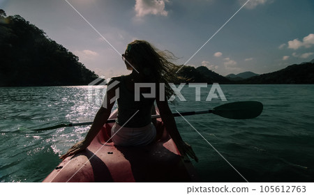 Silhouette of woman tourist raising hands while sailing in canoe along sea bay water to distant hilly island backside view. 105612763