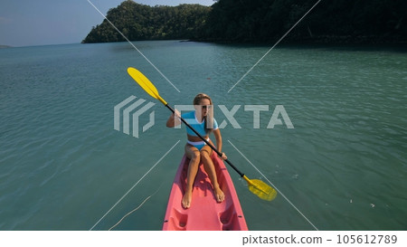 Pretty sportive blonde lady in stylish swimsuit sails on pink plastic canoe along azure ocean bay at exotic resort. Pretty sportive blonde lady in stylish swimsuit sails on pink plastic canoe along azure ocean bay at exotic resort. 105612789