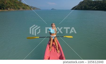 Pretty sportive blonde lady in stylish swimsuit sails on pink plastic canoe along azure ocean bay at exotic resort. Pretty sportive blonde lady in stylish swimsuit sails on pink plastic canoe along azure ocean bay at exotic resort. 105612840