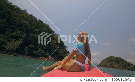 Pretty young blonde woman in stylish swimsuit and sunglasses sits on pink canoe sailing along sea against hills. Pretty young blonde woman in stylish swimsuit and sunglasses sits on pink canoe sailing along sea against hills. 105612846