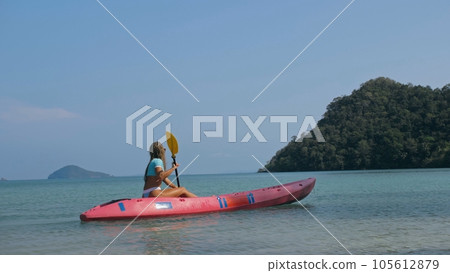 Young blonde woman in blue swimsuit rows pink plastic canoe along azure sea bay past island with palms under blue sky at resort. Young blonde woman in blue swimsuit rows pink plastic canoe along azure sea bay past island with palms under blue sky at resort. 105612879