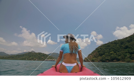Sexy young woman in blue swimsuit and hat swings feet in water on pink canoe on sea against hills backside view. 105612909