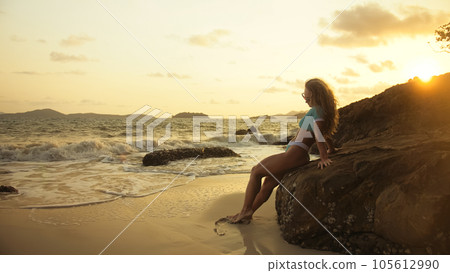 Woman leaning on a rock reef enjoying the warm golden sunset. Wo 105612990