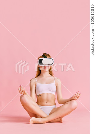 Meditating. Sportive young girl practicing yoga in lotus pose in VR headset glasses. Fitness and lifestyle of future. Meditating. Sportive young girl practicing yoga in lotus pose in VR headset glasses. Fitness and lifestyle of future. 105615819