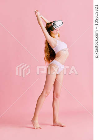 Full-lenght image of young woman in white underwear posing in VR headset glasses over pink background. Full-lenght image of young woman in white underwear posing in VR headset glasses over pink background. 105615821