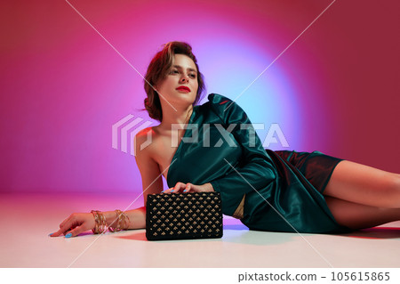 Horizontal image of pretty woman, high fashion model wearing trendy dress with accessories lying on floor over violet-pink neon background. 105615865
