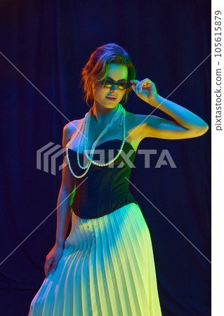 Portrait of woman, young fashion model in fashionable outfit touching sunglasses posing over dark violet background in neon light Portrait of woman, young fashion model in fashionable outfit touching sunglasses posing over dark violet background in neon light 105615879