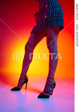 Slim legs in heels of young woman black plaid mini skirt over gradient yellow-red neon background. 105615890