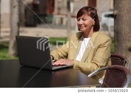 Elegant adult businesswoman works and type at laptop, making call on smartphone in an outdoor cafe at city. Concept of remote work communication from public place, digital freelance, modern lifestyle. 105615920