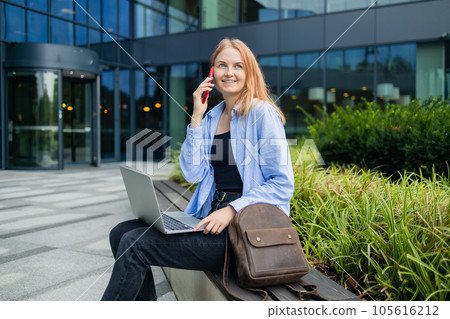 Happy Caucasian student freelancer blonde woman rest use laptop pc computer talk by mobile cell phone look, sitting outdoors against blurred city building. She is keyboarding on laptop. Freelance work 105616212