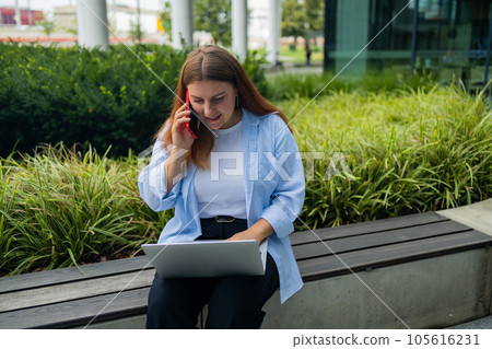 Sad Caucasian student freelancer 30s woman rest use laptop pc computer talk by mobile cell phone look, outdoors sits at city cafe against blurred city building. She is keyboarding on laptop. Freelance 105616231