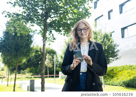 Smiling young Caucasian business woman leader entrepreneur, professional manager holding smartphone using software applications standing on the street in big city on sky background. Smiling young Caucasian business woman leader entrepreneur, professional manager holding smartphone using software applications standing on the street in big city on sky background. 105616239