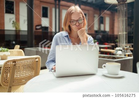 Stressed young blonde woman using laptop at cafe, sitting at table with notepad and cup of coffee alone Stressed young blonde woman using laptop at cafe, sitting at table with notepad and cup of coffee alone 105616275