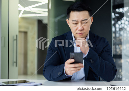 Mature thinking asian boss inside office workplace with phone in hands, successful businessman thinking sitting with laptop in business clothes. Mature thinking asian boss inside office workplace with phone in hands, successful businessman thinking sitting with laptop in business clothes. 105616612
