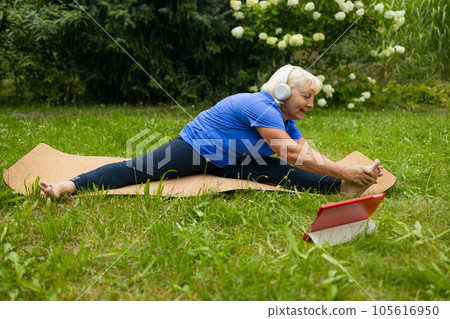 Athletic 50s senior woman uses digital tablet during workout watches online session or workout program poses at yoga mat outdoors dressed in sport clothes alone in the park or forest in nature  105616950
