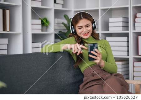 A beautiful and cute Asian woman wearing headphones sits on a desk and uses a smartphone to listen to music, happy and relaxed listening to music at the home A beautiful and cute Asian woman wearing headphones sits on a desk and uses a smartphone to listen to music, happy and relaxed listening to music at the home 105620527