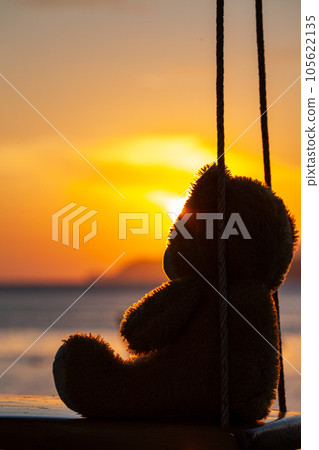 lonely Teddy Bear sitting on a swing in front Rawai beach to see sunrise. scenic reflection of beautiful sunshine above fishing boats are floating in the Rawai sea. beautiful golden sky background. lonely Teddy Bear sitting on a swing in front Rawai beach to see sunrise. scenic reflection of beautiful sunshine above fishing boats are floating in the Rawai sea. beautiful golden sky background. 105622135