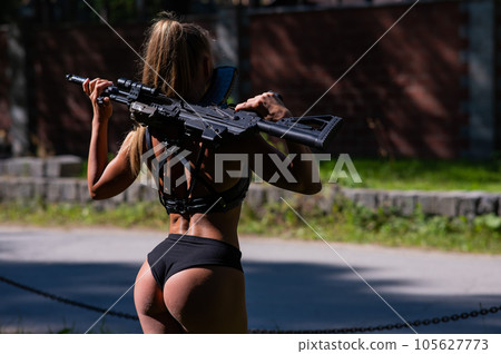 A caucasian woman in a bikini holds a machine gun against a brick wall. 105627773