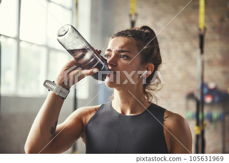 Close up of young athletic woman in earphones is drinking water while exercising in gym. 105631969
