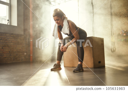 Hard workout. Tired athletic woman with perfect body in sports wear wearing towel on her shoulders while sitting at gym after workout Hard workout. Tired athletic woman with perfect body in sports wear wearing towel on her shoulders while sitting at gym after workout 105631983