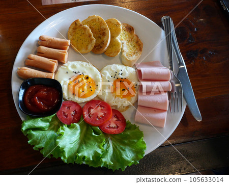 Two fried egg with Bread with sausage and ham in white circle plate, Breakfast with tomato green color vegetables on brown table 105633014