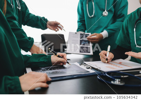 Medical team having a meeting with doctors in white lab coats and surgical scrubs seated at a table discussing a patients working online using computers in the medical industry. 105634342