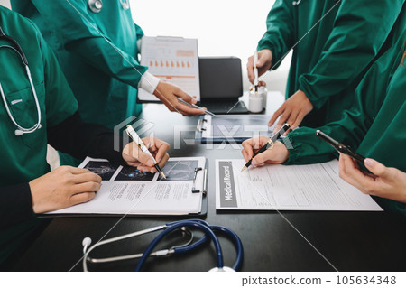Medical team having a meeting with doctors in white lab coats and surgical scrubs seated at a table discussing a patients working online using computers in the medical industry. Medical team having a meeting with doctors in white lab coats and surgical scrubs seated at a table discussing a patients working online using computers in the medical industry. 105634348