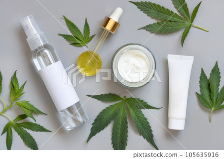 Cosmetic bottles, jars and pipette with blank label near green cannabis leaves on grey. Mockup Cosmetic bottles, jars and pipette with blank label near green cannabis leaves on grey. Mockup 105635916