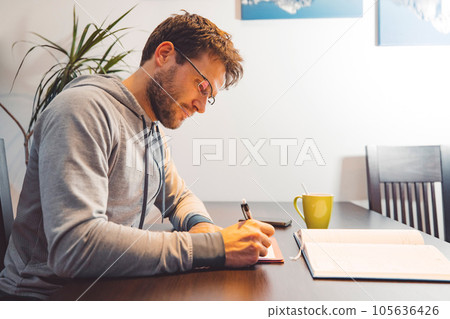 Side view of man with glasses wearing a tracksuit, sitting by the table making notes studying at home 105636426
