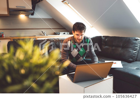 Bearded adult man sitting on black leather sofa studying online on his laptop at home 105636427