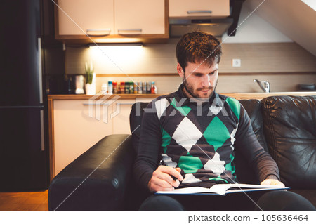 Waist up adult man in green and black sweater sitting on a leather sofa studying for exam at home Waist up adult man in green and black sweater sitting on a leather sofa studying for exam at home 105636464