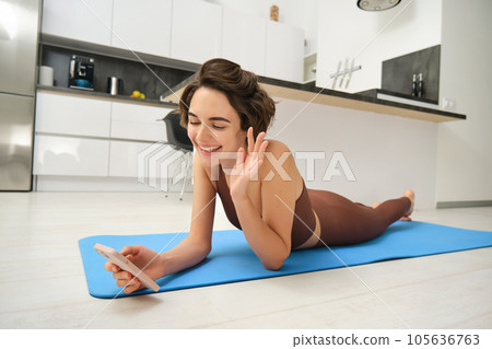 Portrait of fitness girl video chats on smartphone app, saying hello at mobile phone camera, connecting to online workout from home, lying on rubber yoga mat 105636763