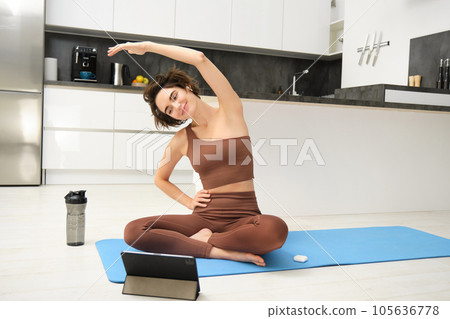 Young woman looking at digital tablet, connects to stretching fitness online class, workout, sits on yoga rubber mat at home, follows gym instructor exercises remotely 105636778