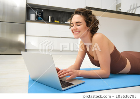 Portrait of beautiful sportswoman, fitness girl watching videos on laptop during workout, follow online pilates videos while exercise at home, wearing activewear in kitchen 105636795