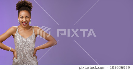 Cheeky good-looking african-american woman in silver glittering dress hold hand waist show tongue daring winking camera feel lucky standing thrilled dancing have fun partying, blue background 105636959