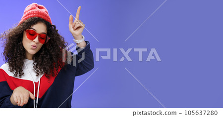 Cute party girl having fun making disco movements folding lips as enjoying cool song dancing joyfully wearing stylish red sunglasses and warm hat looking down enjoying music at party over blue wall Cute party girl having fun making disco movements folding lips as enjoying cool song dancing joyfully wearing stylish red sunglasses and warm hat looking down enjoying music at party over blue wall 105637280