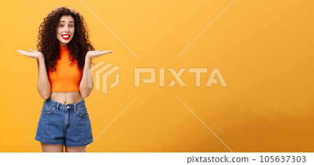 Oops honey sorry. Clueless silly stylish european female with curly hairstyle in cropped top shrugging with hands spread aside in unaware gesture smiling guilty and questioned over orange background 105637303