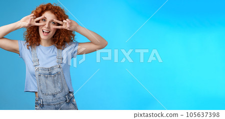 Cheerful cute redhead ginger girl curly haircut show positivity peace victory signs eyes smiling broadly have fun playful silly childish mood wear denim summer overalls blue background 105637398