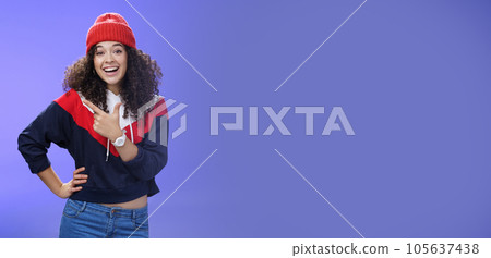 Stylish and sociable friendly-looking attractive woman with curly hair in warm beanie pointing at upper left corner laughing from joy and amazement having fun posing happily over blue background Stylish and sociable friendly-looking attractive woman with curly hair in warm beanie pointing at upper left corner laughing from joy and amazement having fun posing happily over blue background 105637438