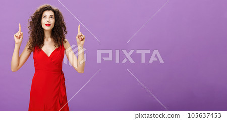 Portrait of intrigued and curious charming young female with curly hairstyle in red stylish evening dress looking and pointing up smiling interested and delighted posing over purple background Portrait of intrigued and curious charming young female with curly hairstyle in red stylish evening dress looking and pointing up smiling interested and delighted posing over purple background 105637453