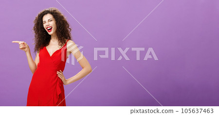 Joyful carefree elegant female with curly hairstyle having fun on awesome party in red luxurious dress holding hand on waist winking and smiling broadly pointing left, standing over purple background Joyful carefree elegant female with curly hairstyle having fun on awesome party in red luxurious dress holding hand on waist winking and smiling broadly pointing left, standing over purple background 105637463