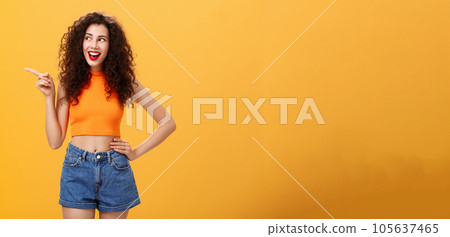 Portrait of delighted enthusiastic stylish young woman with curly hairstyle in red lipstick and cropped top holding hand on waist pointing and looking left amused and happy over orange background Portrait of delighted enthusiastic stylish young woman with curly hairstyle in red lipstick and cropped top holding hand on waist pointing and looking left amused and happy over orange background 105637465